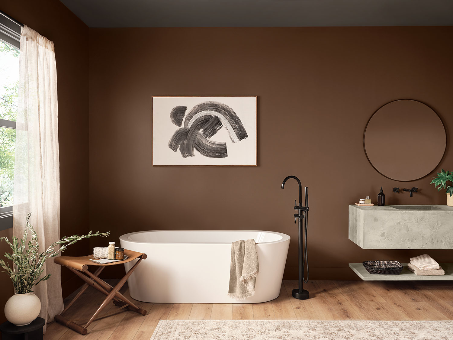 Full room image of bathroom painted Java SW 6090 with stand alone white bathtub, floating modern concrete sink, circle mirror, black and white artwork, leather folding stool holding bath products and two plants