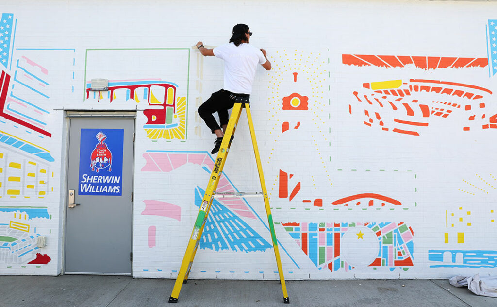 man sitting on a ladder while painting a mural outside of a Sherwin-Williams paint store.