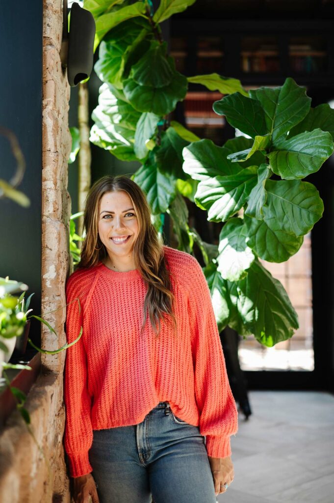 Woman in coral sweater standing in front of large fiddle leaf fig tree.