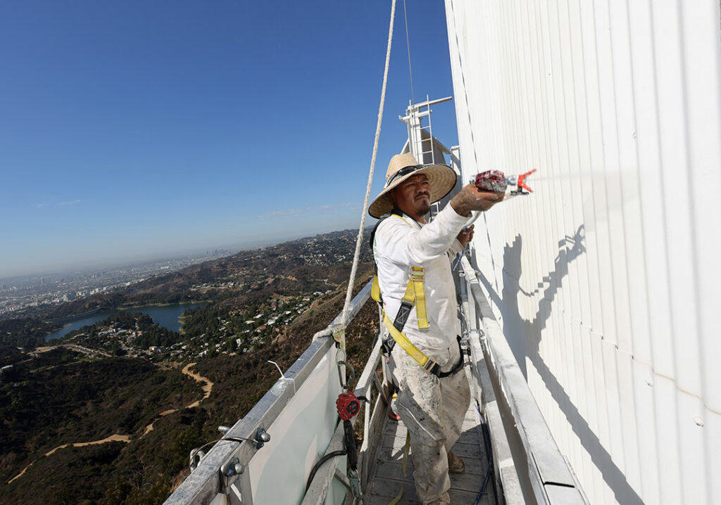 Man on scaffolding painting the Hollywood sign with LA in background.