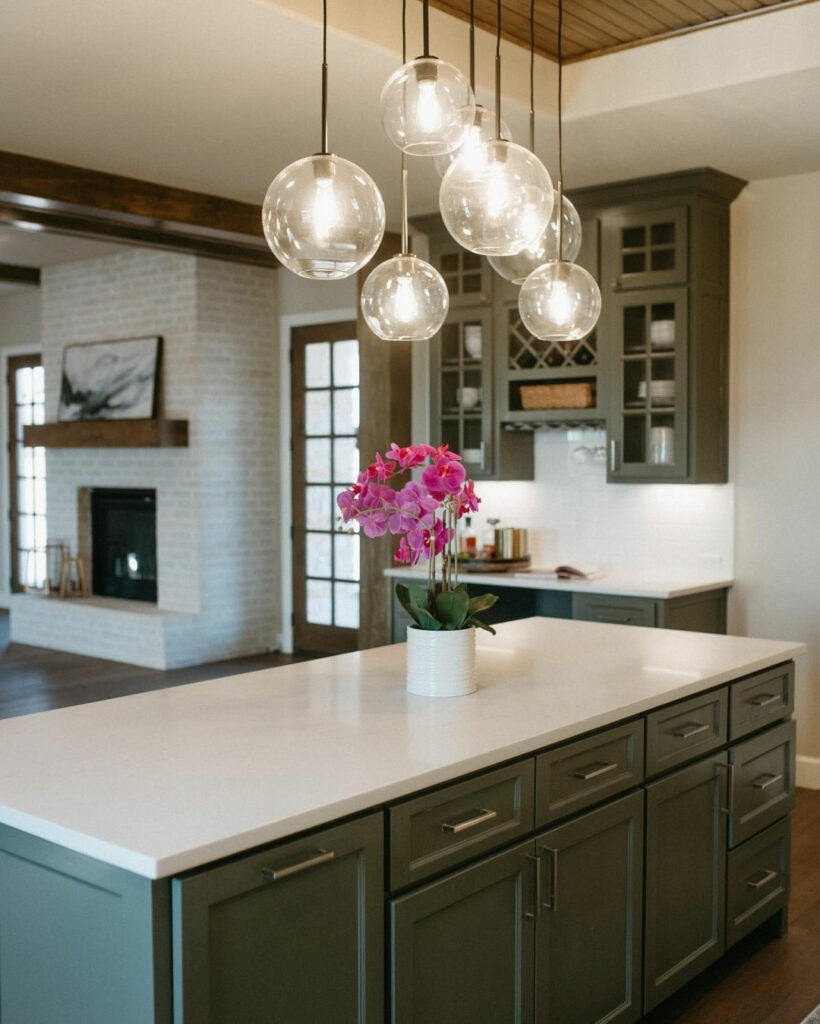 Open-concept country kitchen with a free-standing island painted in a charcoal gray color called Night Owl SW 7061 from Sherwin-Williams. Hanging glass globe light fixtures illuminate the space. Walls in the background are painted in Accessible Beige SW 7036.