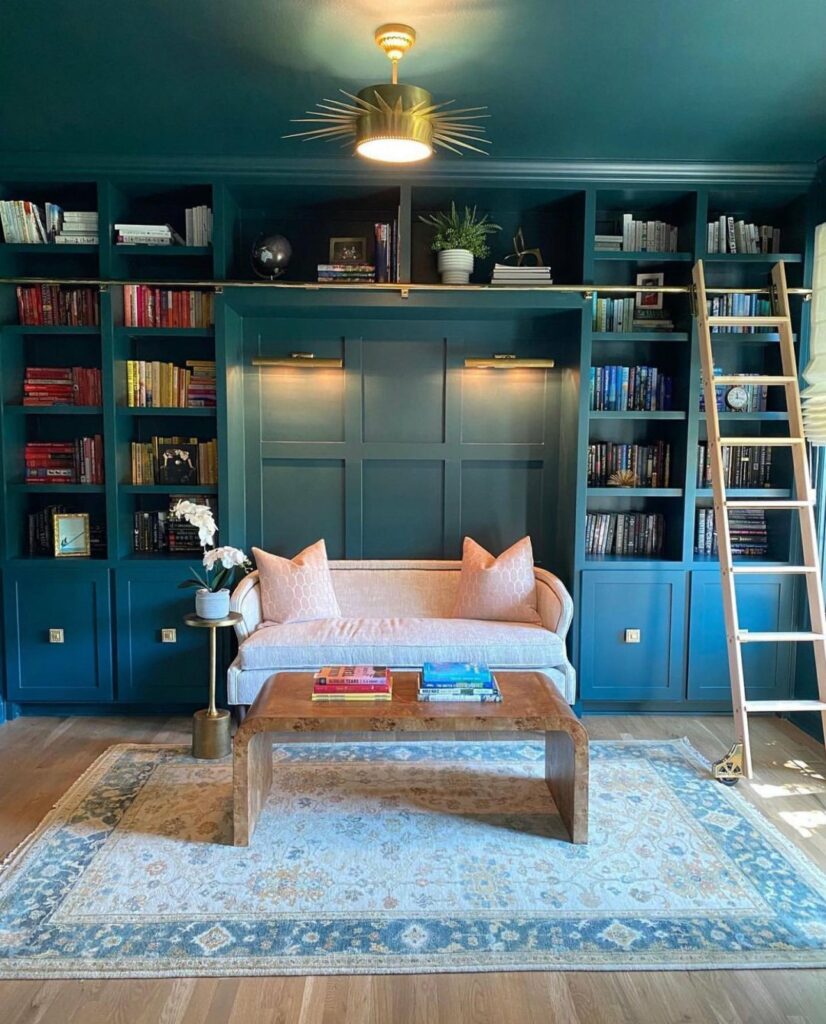 Home library with shelves and ceiling painted in a deep, aqua blue color called Cascades SW 7623 from Sherwin-Williams. In the middle of the room is a soft pink sofa and a curved wood coffee table. A wooden, sliding ladder is attached to the shelves. Lighting comes from a side window as well as a starburst light fixture on the ceiling.