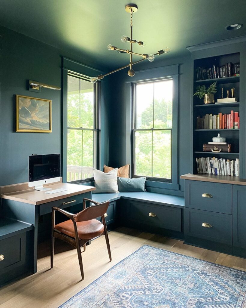 Monochromatic home office with walls, window trim, cabinets, and ceiling painted in a deep, dramatic blue-green color from Sherwin-Williams called Mount Etna SW 7625. Natural sunlight from two corner windows illuminates the space adding shadowed drama.