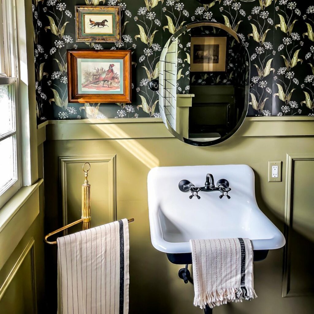Retro bathroom with a white porcelain sink, floral wallpaper, an oval mirror, and wood wainscotting painted in an ochre yellow/green color from Sherwin-Williams called Messenger Bag SW 7740. A window on the left of the room lets natural sunlight stream in, creating beautiful shadows on the wall.