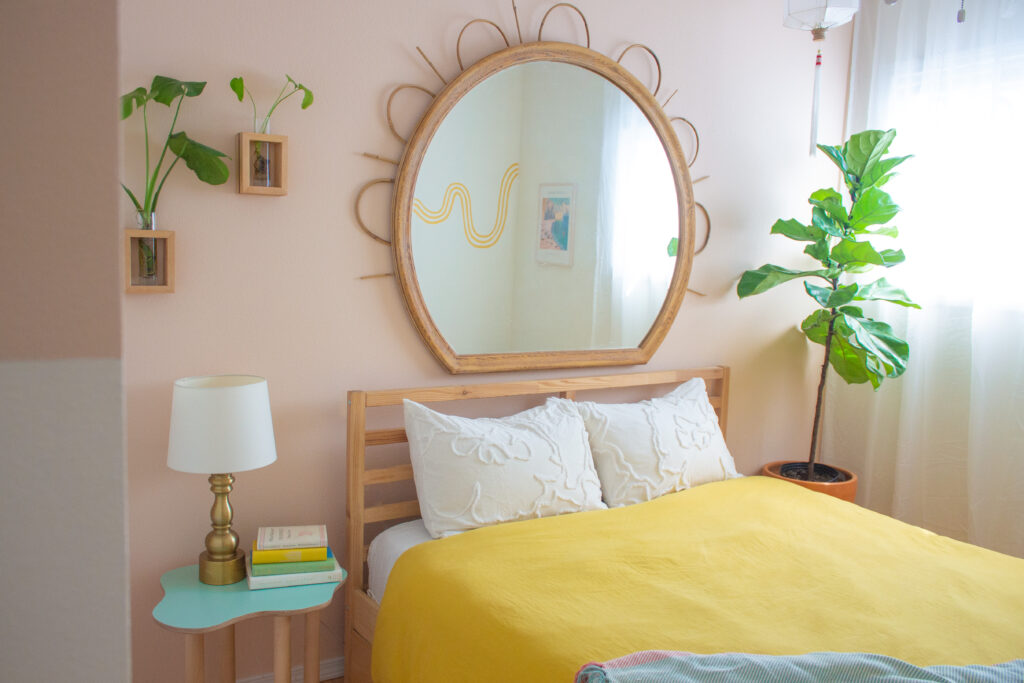 A bright, naturally lit bedroom with brightly colored, yellow bedspread and mirror hanging over wood headboard.