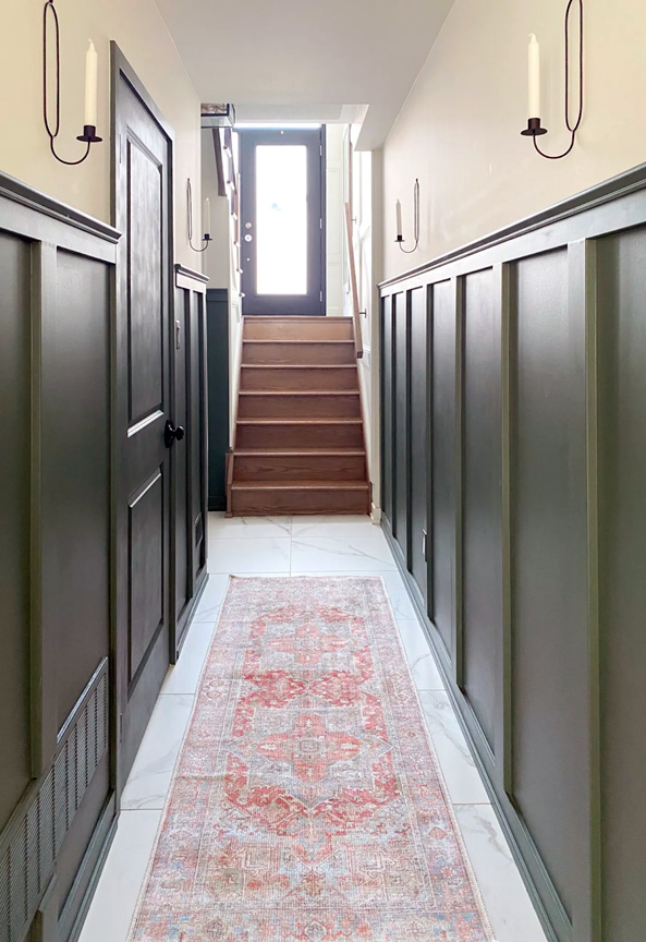 A long, narrow hallway filled with natural light and covered halfway up with wainscoting painted in SW 6208 Pewter Green.